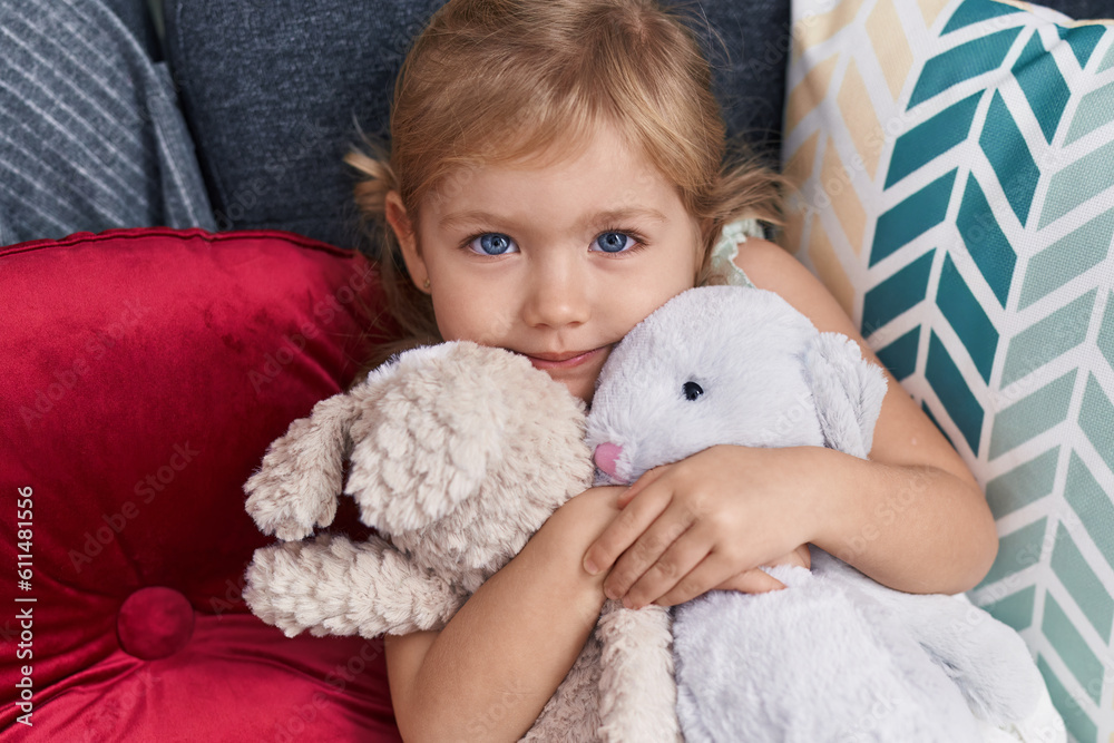 Adorable blonde girl hugging dolls lying on sofa at home