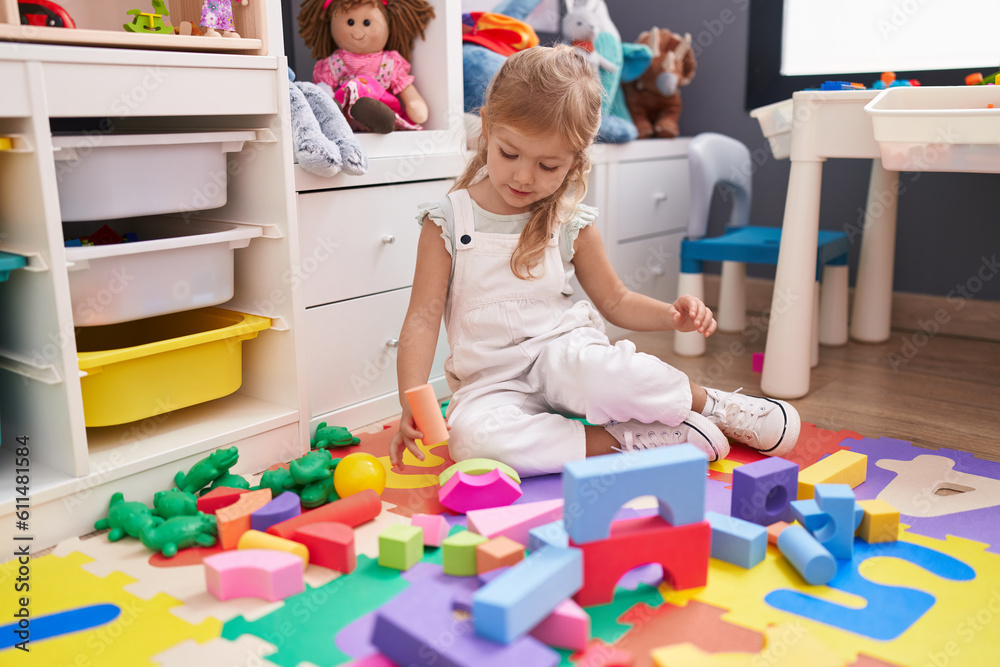 Fototapeta premium Adorable blonde girl playing with construction blocks sitting on floor at kindergarten