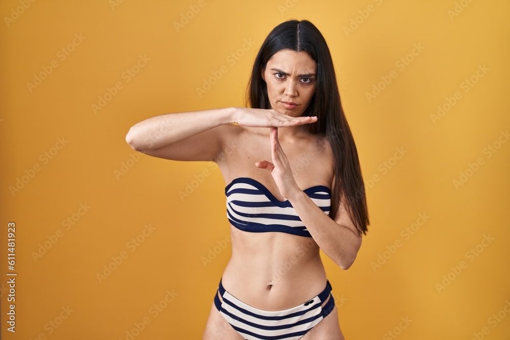 Young brunette woman wearing bikini over yellow background doing time out gesture with hands, frustrated and serious face Young brunette woman wearing bikini over yellow background doing time out gesture with hands, frustrated and serious face
