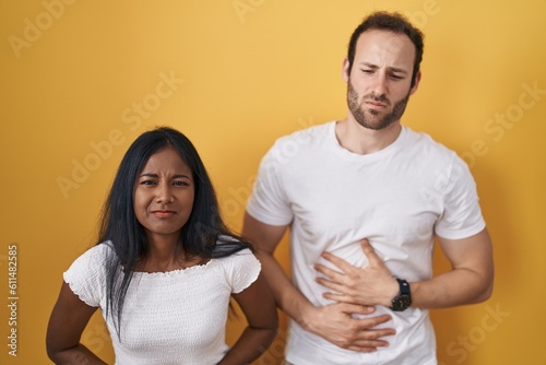 Interracial couple standing over yellow background with hand on stomach because indigestion, painful illness feeling unwell. ache concept.
