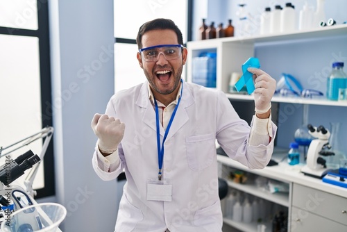 Young hispanic man with beard working at scientist laboratory holding blue ribbon screaming proud  celebrating victory and success very excited with raised arms