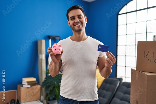 Young hispanic man with beard holding piggy bank and credit card at new home smiling with a happy and cool smile on face. showing teeth.