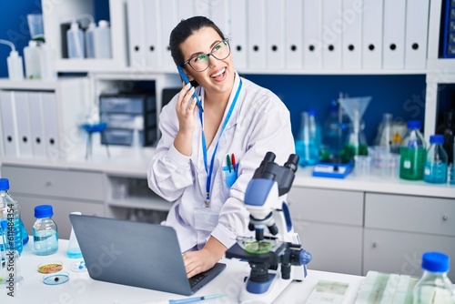 Young hispanic woman scientist talking on smartphone using laptop at laboratory