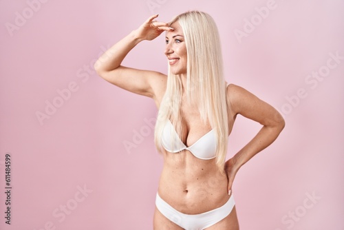Caucasian woman wearing lingerie over pink background very happy and smiling looking far away with hand over head. searching concept.