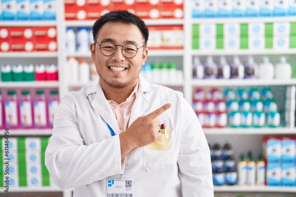 Fototapeta premium Chinese young man working at pharmacy drugstore cheerful with a smile on face pointing with hand and finger up to the side with happy and natural expression