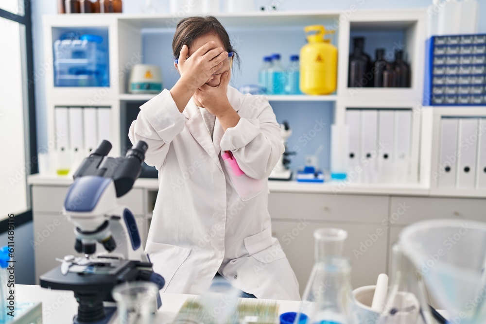 Hispanic girl with down syndrome working at scientist laboratory covering eyes and mouth with hands, surprised and shocked. hiding emotion Hispanic girl with down syndrome working at scientist laboratory covering eyes and mouth with hands, surprised and shocked. hiding emotion