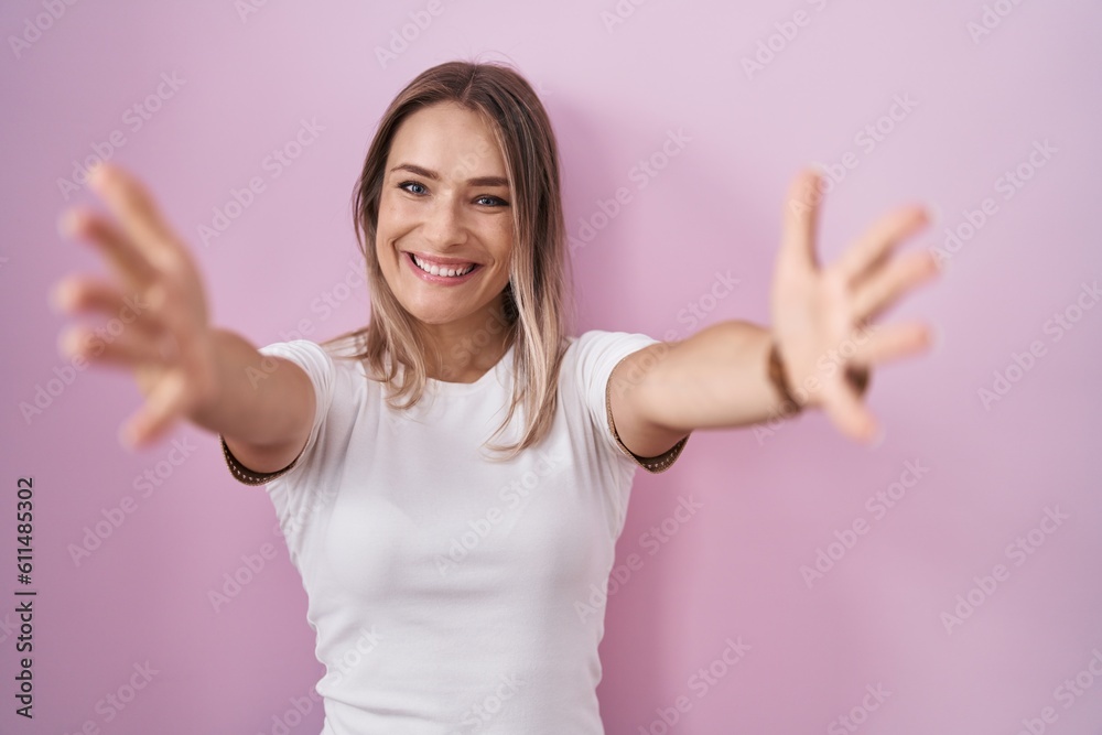 Fototapeta premium Blonde caucasian woman standing over pink background looking at the camera smiling with open arms for hug. cheerful expression embracing happiness.