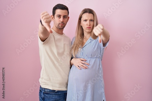 Young couple expecting a baby standing over pink background looking unhappy and angry showing rejection and negative with thumbs down gesture. bad expression.