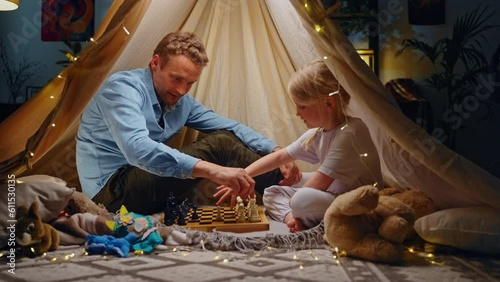Slow motion. Happy father teaches her daughter to play chess, sitting together in a wigwam tent photo