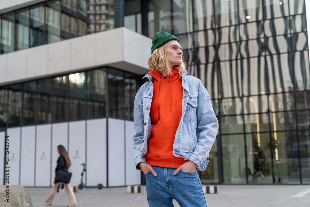 Portrait oeuropean young hipster man, hands in jeans pockets, looking sideways and standing next modern business center in big city. Stylish blond male in denim near shopping malll. Urban lifestyle