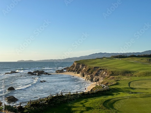 Scenic coastal cliffs with waves crashing against rocky shoreline, expansive green golf course stretching toward the horizon under a clear blue sky.