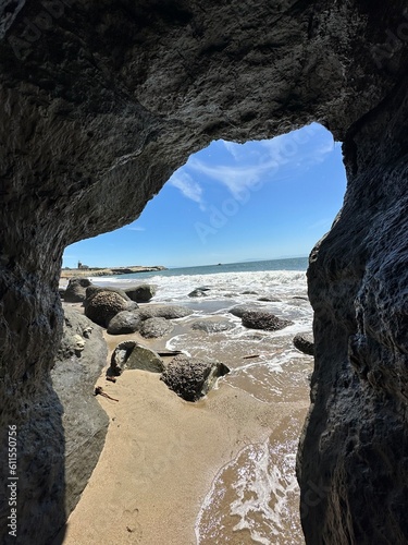 Ocean waves rolling onto sandy beach seen through a rocky cave opening, with large boulders scattered along the shoreline under a bright sunny blue sky.
