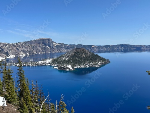 Crater Lake National Park overlook with Wizard Island in the center, surrounded by deep blue water, snowy cliffs, and evergreen trees in Oregon