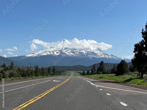 Mount Shasta looming over an empty two-lane highway stretching into the distance with fluffy clouds crowning the snowy peak against a clear blue sky.