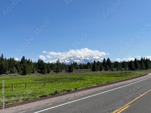 Scenic highway view with Mount Shasta covered in snow and surrounded by clouds, green meadows and pine trees under a bright blue summer sky in Northern California.
