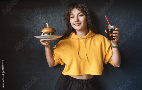 Attractive young woman with curly dark hair in a yellow hoodie holds a burger and coca cola in a glass in her hands on a black background