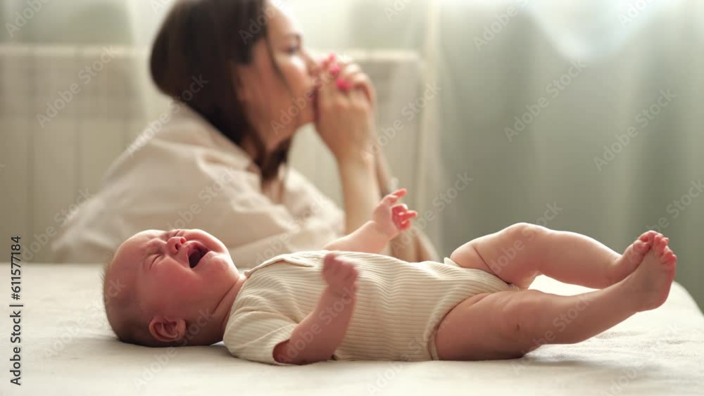 Tired infant girl cries on double bed against depressed mother sitting on floor. Baby daughter wants to eat getting parent attention