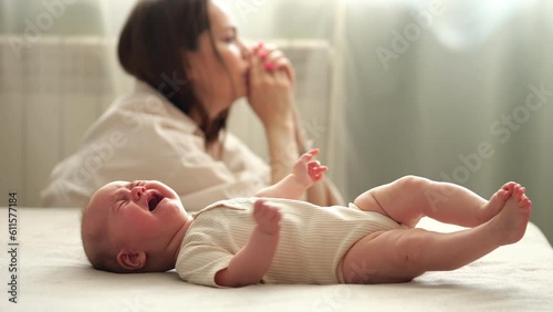 Tired infant girl cries on double bed against depressed mother sitting on floor. Baby daughter wants to eat getting parent attention