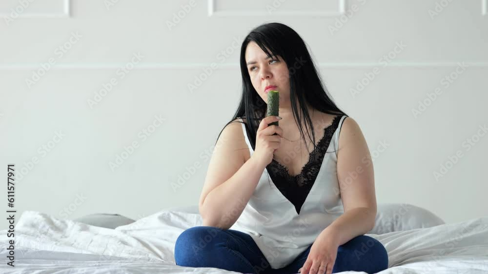 Black-haired woman eats cucumber with disgusted and exhausted expression. Overweight lady looks tired from having healthy diet at home Black-haired woman eats cucumber with disgusted and exhausted expression. Overweight lady looks tired from having healthy diet at home