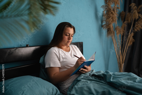 Cute young woman writes a diary while lying in bed in the morning. Rest, sleeping, comfort and people