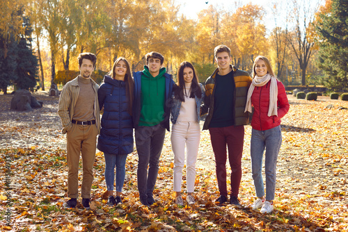 Full length portrait of a group of happy friends walking in the autumn park and hugging. Young smiling caucasian people standing and looking at camera outdoors. Friendship and togetherness concept.