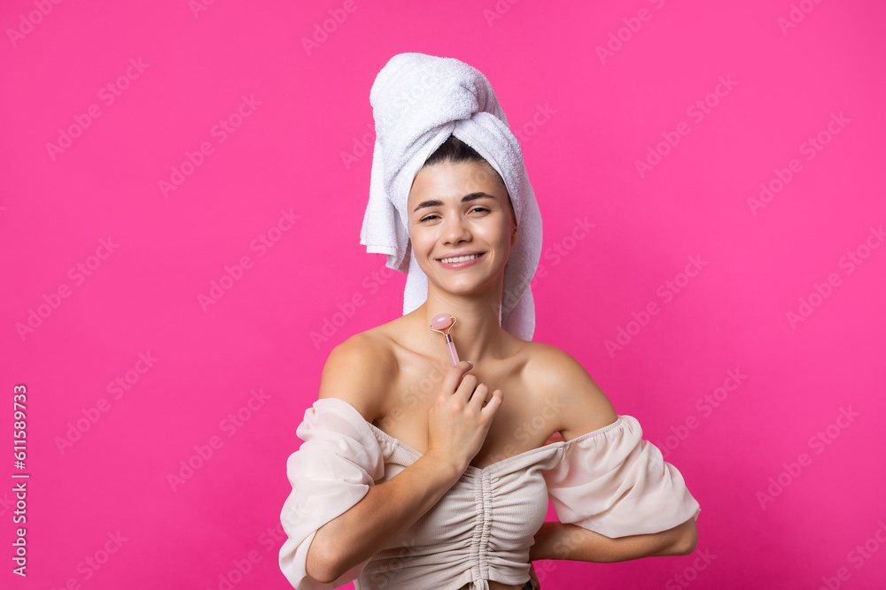 A beautiful cheerful attractive girl with a towel on her head holds a cosmetic roller near her face against a pink background. A beautiful cheerful attractive girl with a towel on her head holds a cosmetic roller near her face against a pink background.