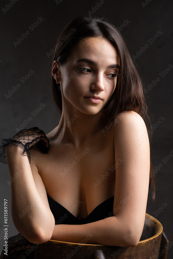 Classic dark studio portrait of a young brunette woman in black clothes who is sitting on a chair. Classic dark studio portrait of a young brunette woman in black clothes who is sitting on a chair.