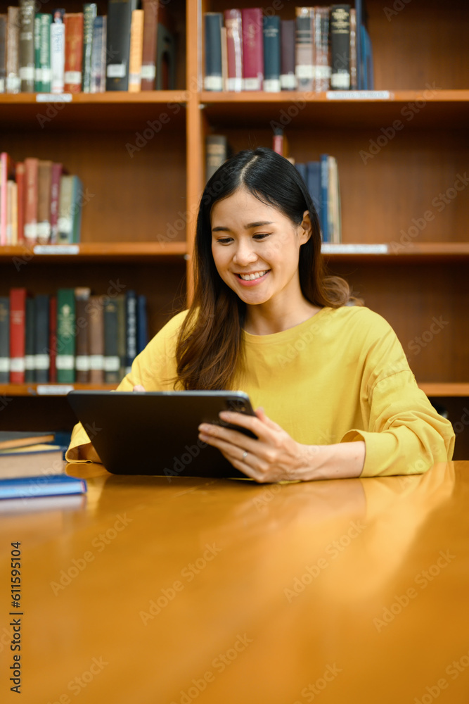 Smiling female university student using digital tablet, doing research, learning lessons, preparing for exams in library Smiling female university student using digital tablet, doing research, learning lessons, preparing for exams in library