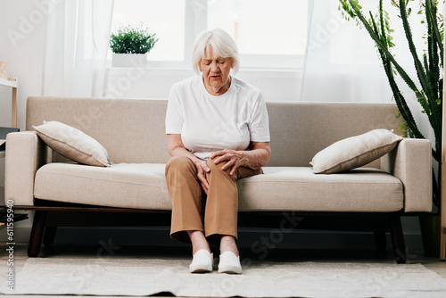 Elderly woman severe pain in her leg sitting on the couch, health problems in old age, poor quality of life. Grandmother with gray hair holds on to her sore knee, problems with joints and ligaments. 