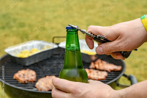 Butelka piwa otwierana otwieraczem na rozmytym tle grilla