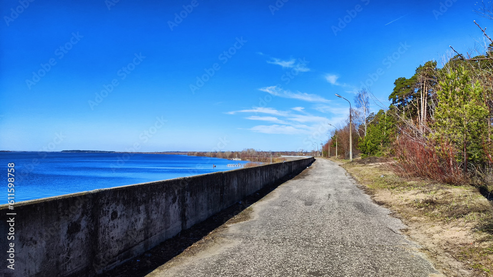 Asphalt path on the embankment near the water of the lake, sea, river, reservoir and blue distance with the sky with white clouds and horizon. Natural landscape with water on a sunny day