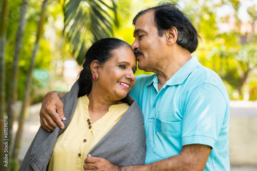 Happy senior husband kissing his wife's forehead by hugging at park - concept of affection, physical intimacy and romantic moment.