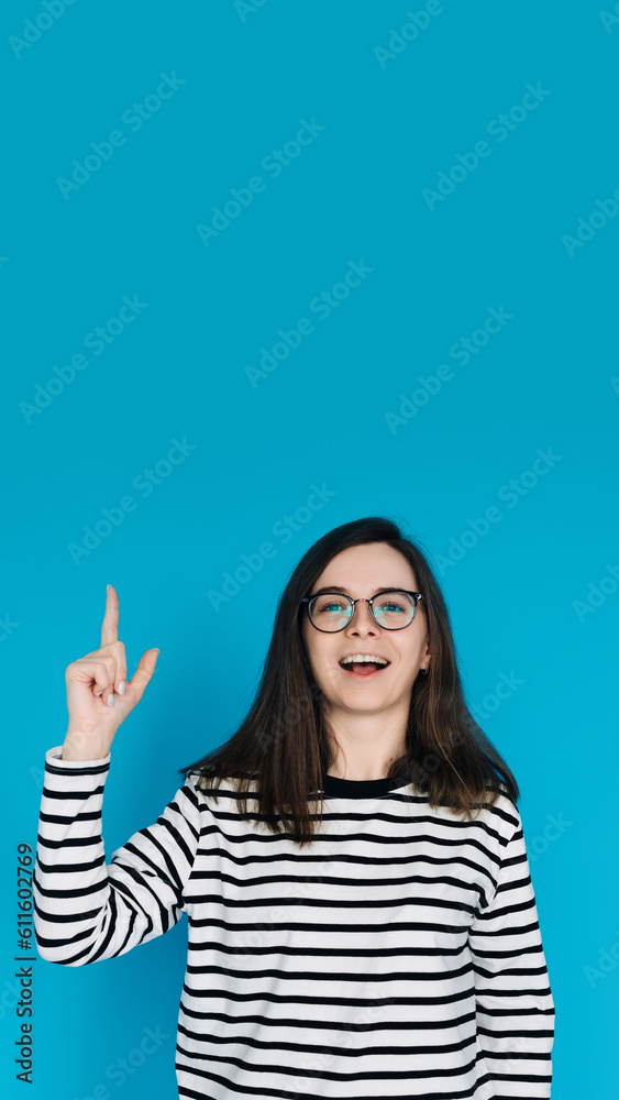 Excited Cheerful Woman in Striped Sweater with Glasses Pointing Up - Energetic Female Model Gesturing towards Empty Space - Isolated on Blue Background - Perfect for Advertising, Promotions Concepts Excited Cheerful Woman in Striped Sweater with Glasses Pointing Up - Energetic Female Model Gesturing towards Empty Space - Isolated on Blue Background - Perfect for Advertising, Promotions Concepts