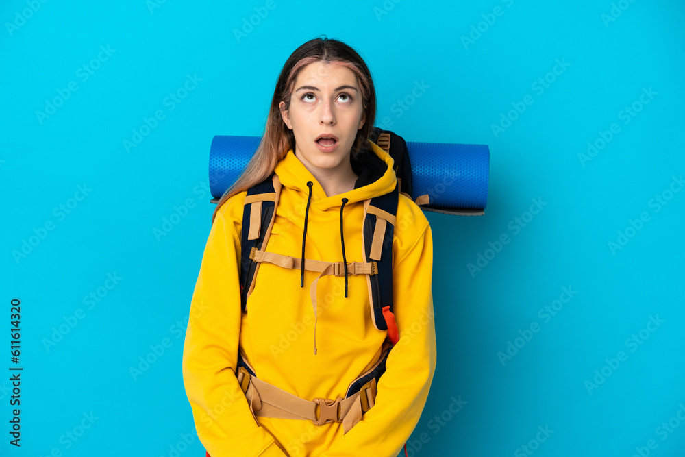 Young mountaineer woman with a big backpack isolated on blue background looking up and with surprised expression Young mountaineer woman with a big backpack isolated on blue background looking up and with surprised expression