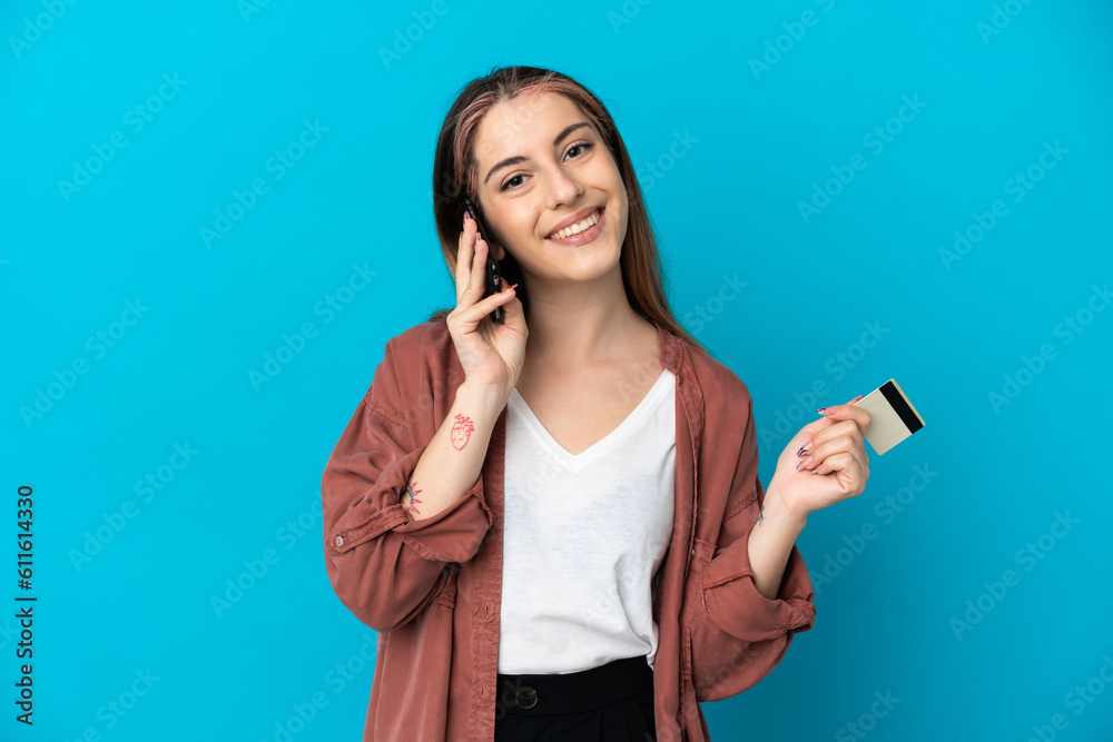Young caucasian woman isolated on blue background keeping a conversation with the mobile phone and holding a credit card Young caucasian woman isolated on blue background keeping a conversation with the mobile phone and holding a credit card