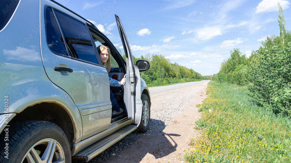 A young girl with blonde hair is sitting in a car with the door open on the side of the road outside the city. The concept of traveling by car. High quality photo