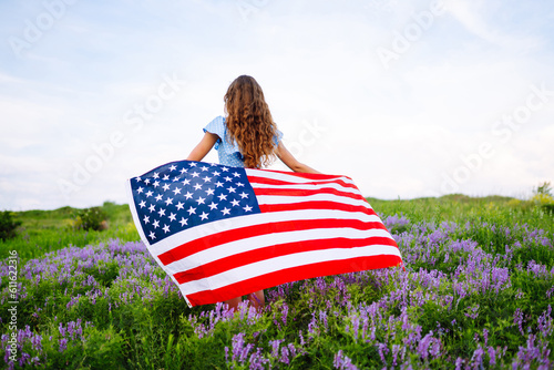 Young woman waving American flag on blooming meadow. 4th of July. Independence Day. Patriotic holiday. USA flag fluttering in the wind.