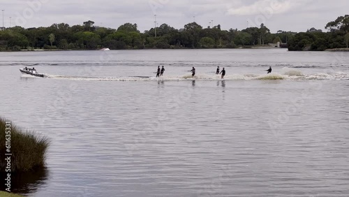 Speedboat tows 6 performance skiers in black wetsuits on Swan River. Tree covered far bank. Light cloud cover