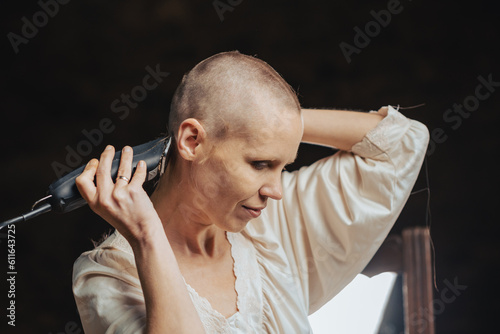 Portrait of a bald woman cutting her hair with a clipper on a dark background.