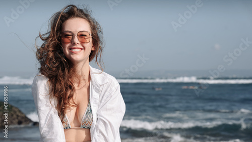 Girl on the beach in a white shirt, bikini and glasses. Looks into the camera and smiles. Ocean coast.