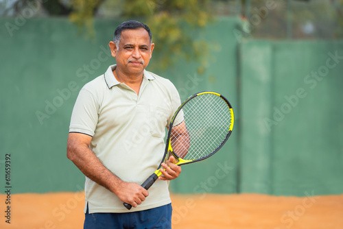 Happy smiling indian senior men with racquet and ball looking camera at tennis court - concept of confident, successful and relaxation