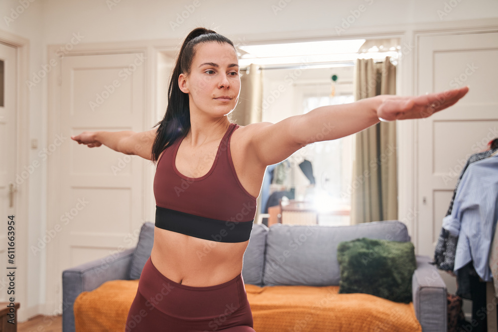 Young calm woman wearing home clothes practicing yoga in warrior pose at the light living room