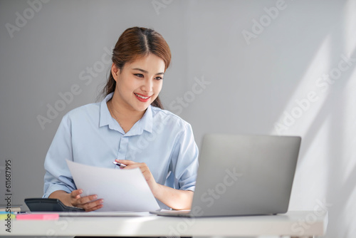 Charming Young asian businesswoman sitting on laptop computer in the office, making report calculating balance Internal Revenue Service checking document.