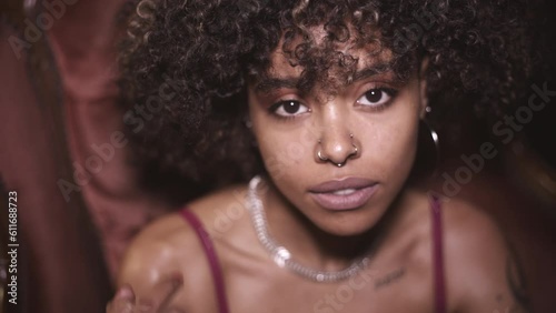 Young afro-haired and tattooed girl sitting in an old-fashioned armchair, giving the middle finger to the camera.