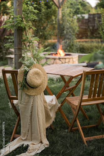 Wooden furniture in the garden against the backdrop of a fire on the patio. Among the branches of the bush stands a wooden table and two wooden chairs. One of them is wearing a straw hat and a plaid.