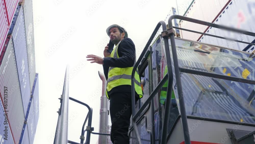 Supervisor engineer or Businessman wear vest and helmet safety talking mobile phone and standing on forklift look around portyard in warehouse area. Logistics and shipping concept Supervisor engineer or Businessman wear vest and helmet safety talking mobile phone and standing on forklift look around portyard in warehouse area. Logistics and shipping concept