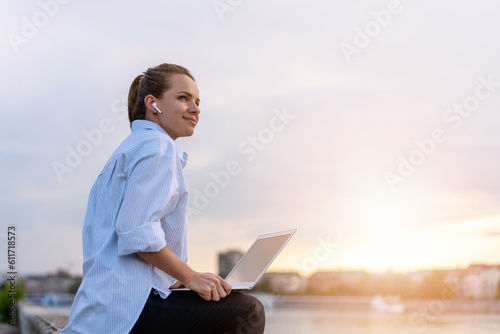 A young city woman is sitting on the waterfront with her laptop enjoying the sunset, outside the office job.
