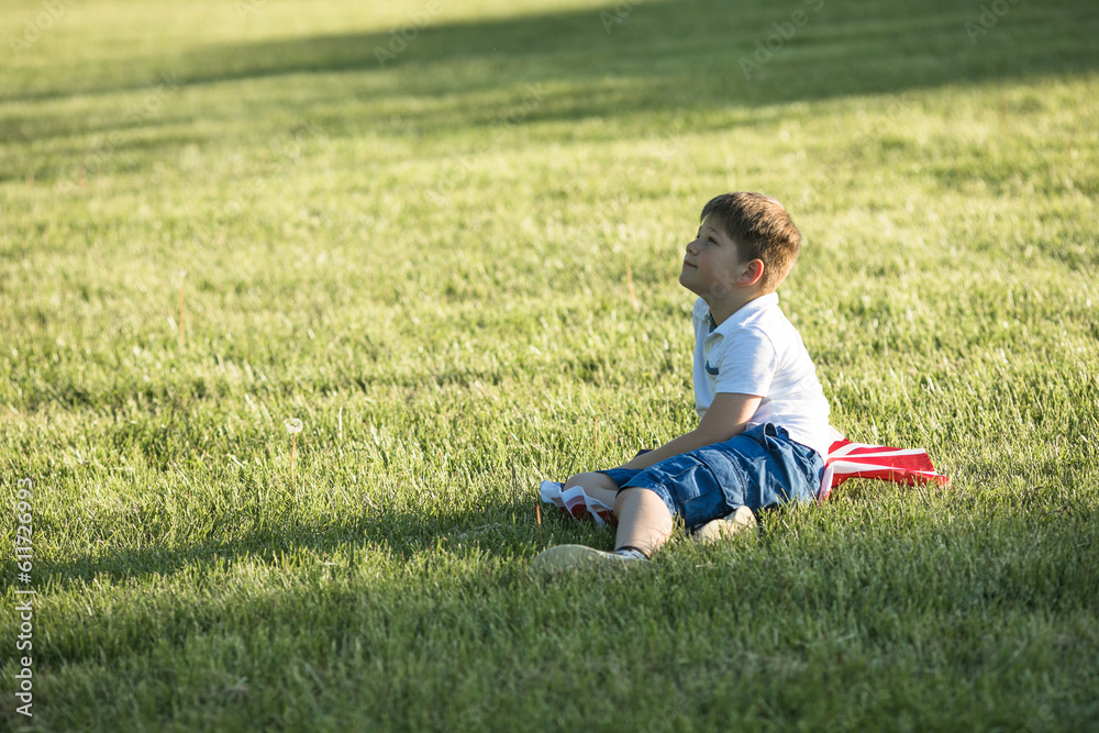 Cute little boy celebrating of July, 4 Independence Day of USA at sunny summer sunset. Child sitting with american flag symbol of United States over field. Proud little american boy Cute little boy celebrating of July, 4 Independence Day of USA at sunny summer sunset. Child sitting with american flag symbol of United States over field. Proud little american boy