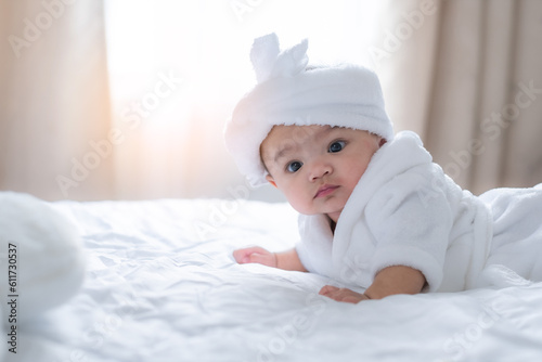 Adorable baby child looking away through camera on white bed, with curtain background and light of rising sun.