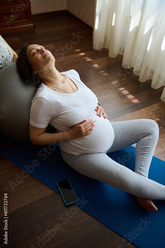 Overhead view beautiful pregnant woman, relaxing with her eyes closed, leaning on a fitball, holding hands on her belly, sitting on a mat at home. Daylight. Prenatal fitnesss and relaxation exercises photo